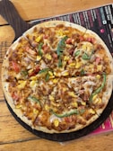 A freshly baked pizza topped with cheese, sliced green bell peppers, corn, and diced tomatoes. The pizza rests on a wooden serving board placed on a wooden table. A colorful menu is partially visible underneath the board.