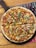 A freshly baked pizza topped with cheese, sliced green bell peppers, corn, and diced tomatoes. The pizza rests on a wooden serving board placed on a wooden table. A colorful menu is partially visible underneath the board.