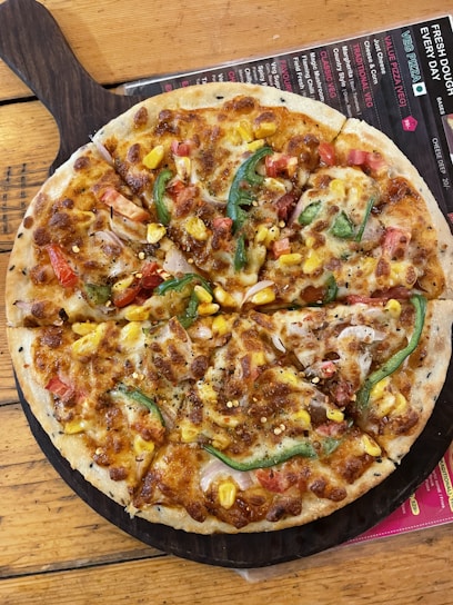 A freshly baked pizza topped with cheese, sliced green bell peppers, corn, and diced tomatoes. The pizza rests on a wooden serving board placed on a wooden table. A colorful menu is partially visible underneath the board.