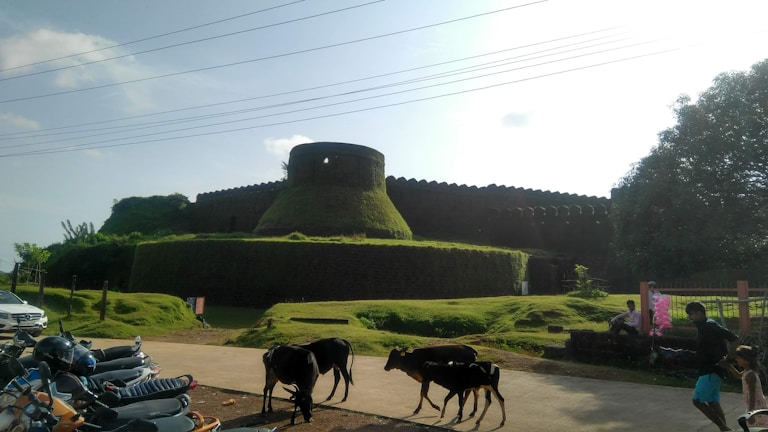 A large, ancient fort with thick, moss-covered walls is surrounded by lush greenery. In the foreground, a few goats walk along a path near parked scooters. There are people on the right side, one of whom appears to be selling cotton candy.