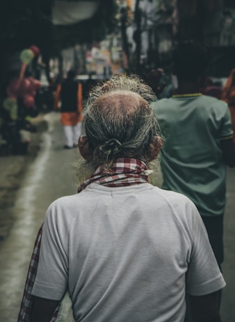 An elderly person with grey hair, viewed from behind, is wearing a white shirt and a red checkered scarf. They are walking on a busy street lined with other people, some wearing colorful clothing. The surroundings are slightly blurred, giving a dynamic and lively atmosphere.