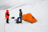 Two people in winter gear stand near an orange tent on a snowy landscape, with climbing equipment and backpacks around them.