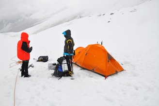 Two people in winter gear stand near an orange tent on a snowy landscape, with climbing equipment and backpacks around them.