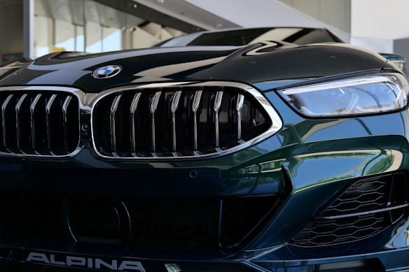 A close-up view of a luxury car's front grille and headlight. The image features sleek, reflective surfaces with intricate design elements, showcasing a modern and sophisticated style.