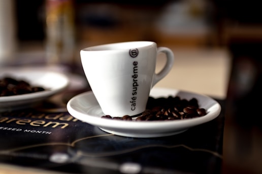 A white coffee cup with the words 'café suprême' printed on its side is placed on a white saucer. The saucer has several coffee beans scattered around it. The background is slightly blurred, giving a cozy and inviting atmosphere.