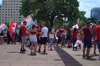 A group of people gather in an outdoor urban setting, holding flags with vibrant colors, possibly participating in a political demonstration or event. Some people are wearing red clothing, and there are tall buildings and trees in the background, suggesting a city square or plaza.