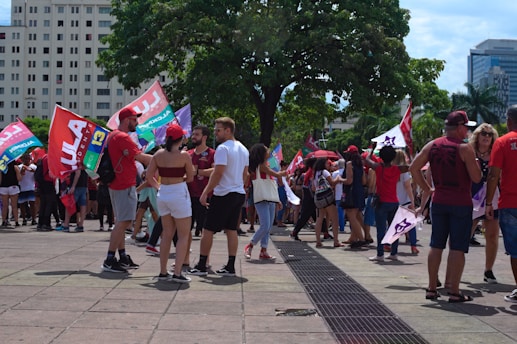 A group of people gather in an outdoor urban setting, holding flags with vibrant colors, possibly participating in a political demonstration or event. Some people are wearing red clothing, and there are tall buildings and trees in the background, suggesting a city square or plaza.