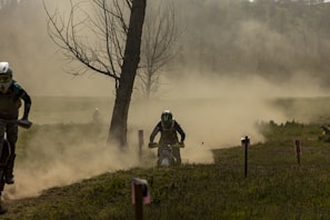 Several motorcyclists are racing through a dusty, off-road track surrounded by sparse trees and foliage. Dirt and dust billow around the riders, creating a hazy background that obscures parts of the landscape. The focus is on the central rider, captured in motion and wearing protective gear.