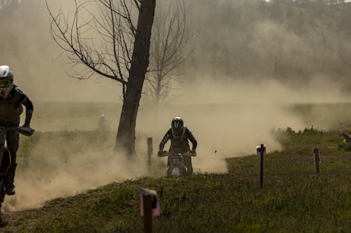 Several motorcyclists are racing through a dusty, off-road track surrounded by sparse trees and foliage. Dirt and dust billow around the riders, creating a hazy background that obscures parts of the landscape. The focus is on the central rider, captured in motion and wearing protective gear.