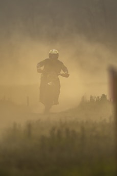 A person on a motorcycle rides through a dusty path, creating a cloud of dust around them. The scene is hazy, and visibility is low due to the dense dust. The rider is wearing a helmet, and their silhouette is highlighted against the dusty background.