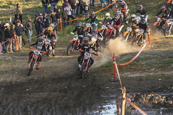 A group of motocross riders racing on a dirt track with spectators watching from the side. Dust is kicked up as the riders maneuver their bikes, each wearing protective gear and helmets. The track is lined with orange barriers, and the crowd is dressed casually, capturing the moment with cameras.