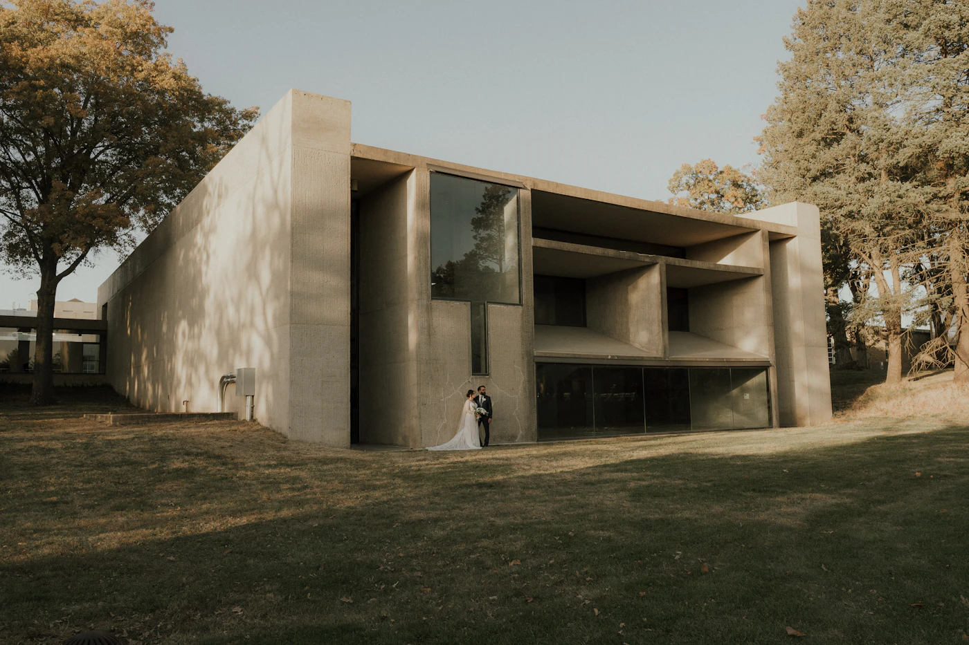 A couple stands small in front of a brutalist concrete house on a sunlit lawn