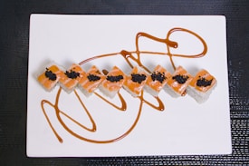 A set of sushi rolls arranged diagonally on a rectangular white plate. The rolls are topped with orange fish, possibly salmon, and small black fish roe. Elegant brown sauce is artistically drizzled on the plate around the rolls, creating an eye-catching presentation.