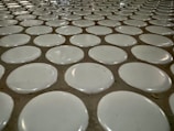 A close-up view of a tiled surface with a repetitive pattern of white circular tiles embedded in a slightly darker grout. The tiles are evenly spaced, creating a uniform and structured appearance.