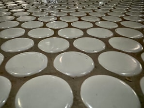 A close-up view of a tiled surface with a repetitive pattern of white circular tiles embedded in a slightly darker grout. The tiles are evenly spaced, creating a uniform and structured appearance.