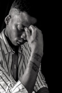 A person sits with their hand placed on their forehead, eyes closed, possibly reflecting or experiencing stress. They wear a striped shirt and a necklace, with visible tattoos on their arm. The image is in black and white, creating a dramatic and contemplative atmosphere.