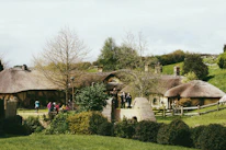A scenic landscape features quaint, rustic houses with thatched roofs surrounded by lush greenery. Several people are seen casually walking, possibly tourists, enjoying the picturesque setting. The scene is tranquil, with a mix of natural and architectural elements.