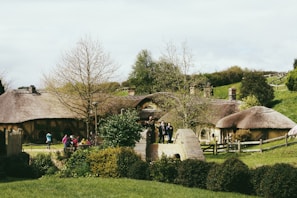 A scenic landscape features quaint, rustic houses with thatched roofs surrounded by lush greenery. Several people are seen casually walking, possibly tourists, enjoying the picturesque setting. The scene is tranquil, with a mix of natural and architectural elements.