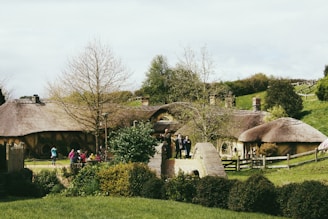 A scenic landscape features quaint, rustic houses with thatched roofs surrounded by lush greenery. Several people are seen casually walking, possibly tourists, enjoying the picturesque setting. The scene is tranquil, with a mix of natural and architectural elements.