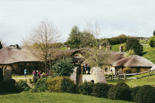 A scenic landscape features quaint, rustic houses with thatched roofs surrounded by lush greenery. Several people are seen casually walking, possibly tourists, enjoying the picturesque setting. The scene is tranquil, with a mix of natural and architectural elements.