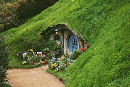 A quaint, round-door hobbit house nestled into a lush, green hillside is adorned with vibrant flowers in an array of clay pots. The entrance is rustic with a charming, cozy feel, surrounded by colorful blossoms and greenery.