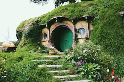 A charming earth-sheltered house with a round door in a vibrant shade of green is nestled within a grassy hill. The entrance is bordered by lush vegetation, blooming flowers, and a rustic stone pathway. A large pumpkin sits on a wooden cart to the left, adding to the quaint and idyllic setting.