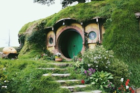 A charming earth-sheltered house with a round door in a vibrant shade of green is nestled within a grassy hill. The entrance is bordered by lush vegetation, blooming flowers, and a rustic stone pathway. A large pumpkin sits on a wooden cart to the left, adding to the quaint and idyllic setting.