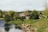 A serene rural scene featuring a quaint cottage with a thatched roof surrounded by lush greenery and trees. A calm body of water borders the foreground, with gentle rolling hills and a rustic fence in the background.