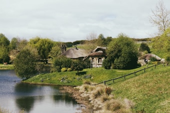 A serene rural scene featuring a quaint cottage with a thatched roof surrounded by lush greenery and trees. A calm body of water borders the foreground, with gentle rolling hills and a rustic fence in the background.