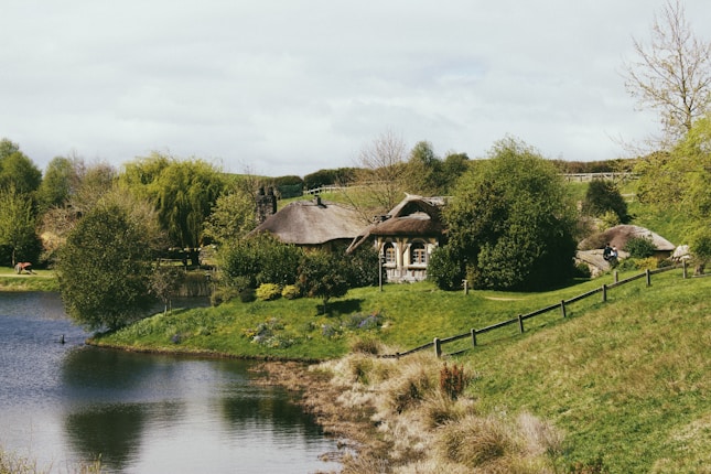 A serene rural scene featuring a quaint cottage with a thatched roof surrounded by lush greenery and trees. A calm body of water borders the foreground, with gentle rolling hills and a rustic fence in the background.