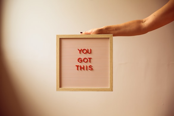 A hand extends from the right, holding a wooden-framed letter board with red letters spelling 'YOU GOT THIS' on a light pink, lined background.