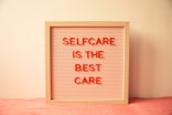 A wooden-framed board with horizontal grooves holds red letters spelling out 'SELFCARE IS THE BEST CARE' against a light pink background. The board is placed against a cream-colored wall with a soft pink cloth underneath.