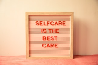 A wooden-framed board with horizontal grooves holds red letters spelling out 'SELFCARE IS THE BEST CARE' against a light pink background. The board is placed against a cream-colored wall with a soft pink cloth underneath.