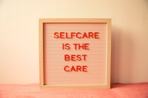A wooden-framed board with horizontal grooves holds red letters spelling out 'SELFCARE IS THE BEST CARE' against a light pink background. The board is placed against a cream-colored wall with a soft pink cloth underneath.