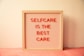 A wooden-framed board with horizontal grooves holds red letters spelling out 'SELFCARE IS THE BEST CARE' against a light pink background. The board is placed against a cream-colored wall with a soft pink cloth underneath.