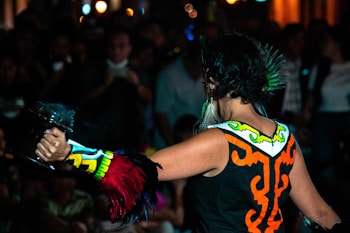 A performer is captured mid-action in a colorful outfit adorned with tribal patterns and vibrant feathers. The setting appears to be a public event with onlookers in the background, creating a lively and dynamic atmosphere.