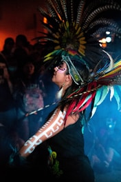 A person is wearing traditional attire with ornate feathered headdress and detailed face paint. Their arm is decorated with white tribal patterns. The atmosphere is dimly lit with a blue hue, and there is a crowd in the background.
