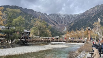 A scenic mountain landscape with a wooden suspension bridge crossing a river. The background features towering mountains partially covered with mist and some snow. The foreground shows the riverbank with gravel and autumn foliage with hues of green and orange. A group of people is walking across the bridge, enjoying the view, and there are more people on the right side taking photos. The overall atmosphere is serene and natural.