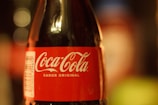 A close-up view of a Coca-Cola bottle with a red label and white text reading 'Coca-Cola Sabor Original'. The bottle is glass and the focus is on the label, with a blurred background.