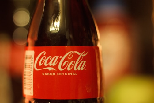 A close-up view of a Coca-Cola bottle with a red label and white text reading 'Coca-Cola Sabor Original'. The bottle is glass and the focus is on the label, with a blurred background.