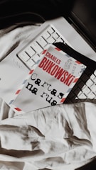A book with the name 'Charles Bukowski' on the cover lies on a laptop keyboard. The cover resembles an airmail envelope with red and blue stripes on the edges. The book is placed over part of the keyboard, and a white fabric is visible underneath.