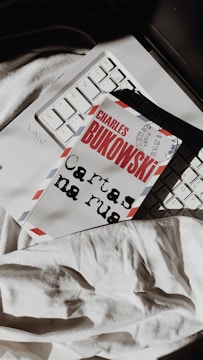 A book with the name 'Charles Bukowski' on the cover lies on a laptop keyboard. The cover resembles an airmail envelope with red and blue stripes on the edges. The book is placed over part of the keyboard, and a white fabric is visible underneath.