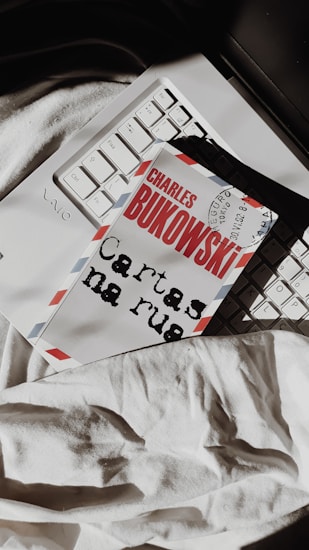 A book with the name 'Charles Bukowski' on the cover lies on a laptop keyboard. The cover resembles an airmail envelope with red and blue stripes on the edges. The book is placed over part of the keyboard, and a white fabric is visible underneath.