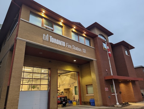 A fire station building with a sign reading 'Toronto Fire Station 132'. The building has a modern architectural design with brick walls and several windows. Bright lights illuminate the entrance and the interior, where a red fire truck is partially visible. A flagpole with a Canadian flag stands beside the entrance.