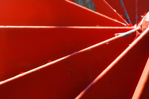 Brightly colored red metal blades arranged in a spiral pattern, showing their surface texture with light and shadow play adding depth to the image.
