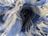 A close-up view of an intertwined fabric with a checkered pattern. The focus is on the intricate folds and frayed edges, creating a dynamic and almost abstract appearance. The fabric features shades of blue and white, contributing to a soft and textured visual.