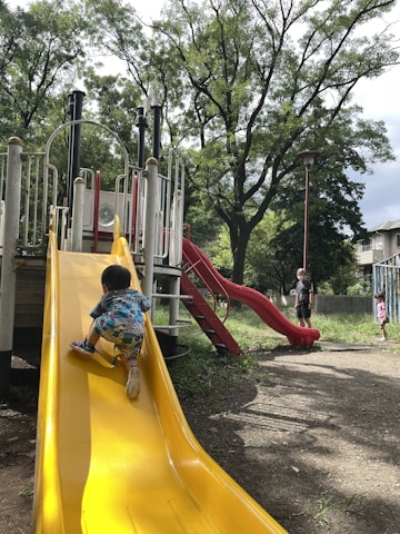 A young child in colorful clothing is climbing up a bright yellow slide in a playground. In the background, there are trees with lush green leaves and another red slide. Two adults and another child can be seen nearby, with one adult watching the child on the slide.