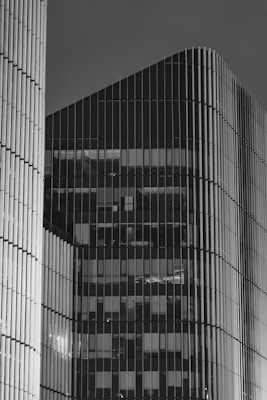 A detailed monochromatic image of modern high-rise buildings with numerous vertical lines and glass panels. The architecture has a sleek, contemporary design featuring smooth curves and reflective surfaces, giving a sense of sophistication and modernity.