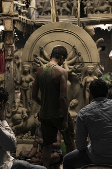 A group of people observing clay sculptures under construction in an artisanal workshop. The sculptures depict human figures and deities, including one with multiple arms. The setting has a rustic and creative atmosphere, filled with partially finished artworks and materials.