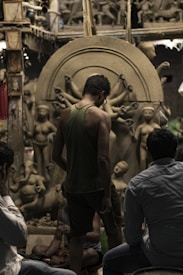 A group of people observing clay sculptures under construction in an artisanal workshop. The sculptures depict human figures and deities, including one with multiple arms. The setting has a rustic and creative atmosphere, filled with partially finished artworks and materials.